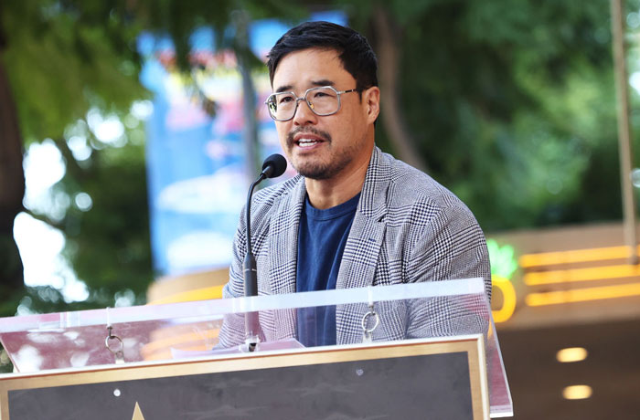 Person speaking at podium outdoors, wearing glasses and a suit jacket, related to Aquaman 2 cast.