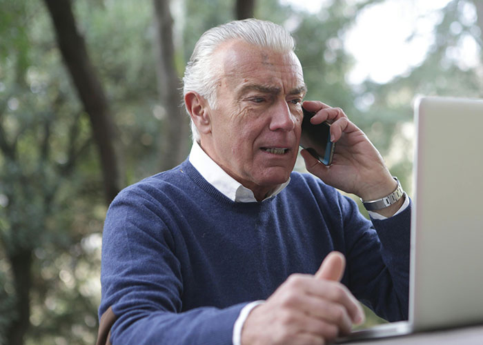 Man talking on phone at laptop, planning to change wedding date, wearing navy sweater in outdoor setting.