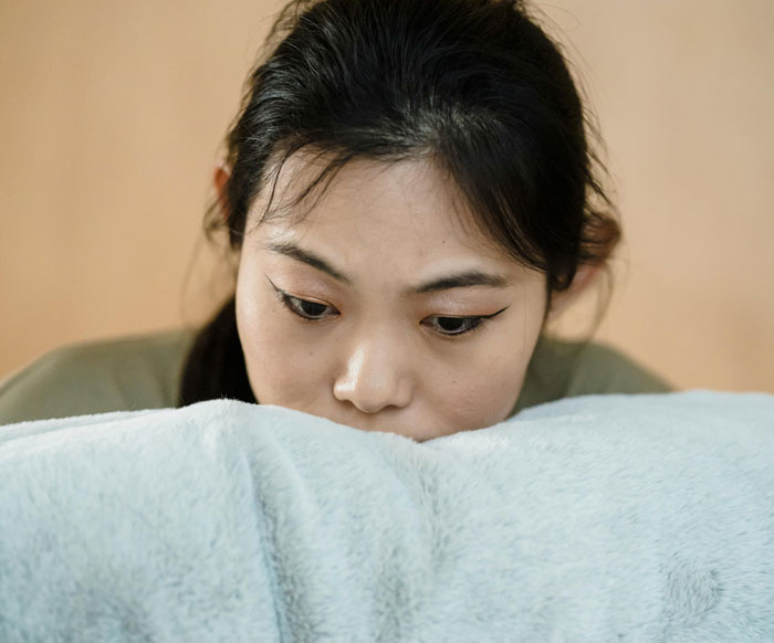 New mom looking pensive in a soft-lit room, resting on a plush cover. Focused expression suggests contemplation.