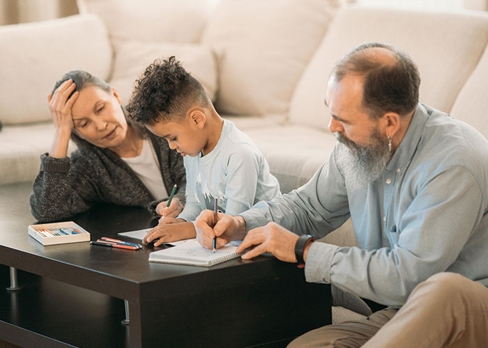 Grandparents and a young boy drawing with colored pencils at a coffee table in a cozy living room.