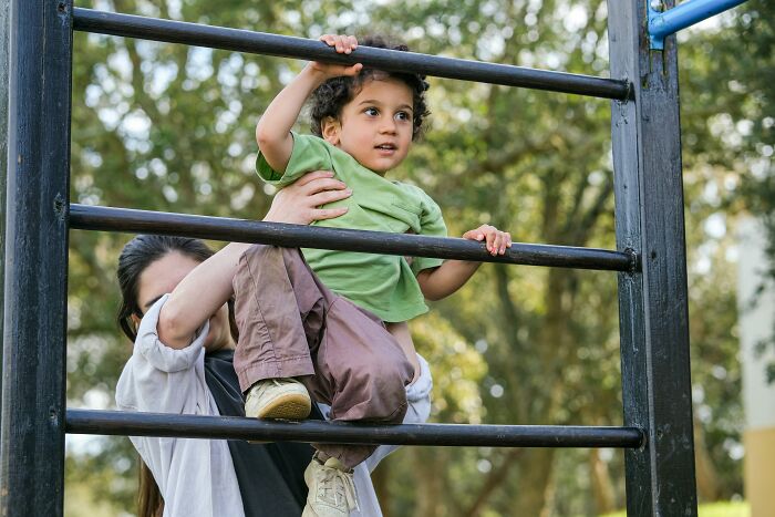 Child climbing playground bars with help from an adult, outdoors setting, representing babysitting moments.