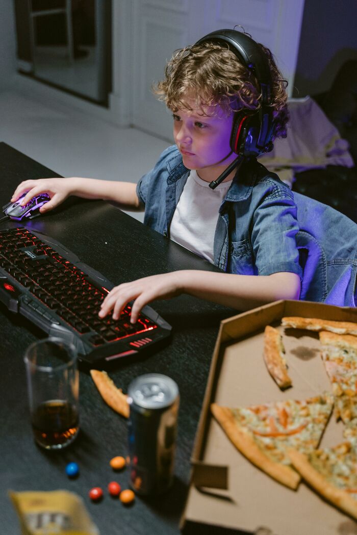 Child wearing a headset playing computer games with a pizza box and snacks on the table, illustrating babysitters' observations.