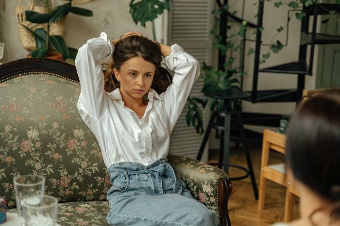 Woman in a white shirt and jeans sitting on a floral couch, surrounded by plants, contemplating bad-good-habit dynamics.