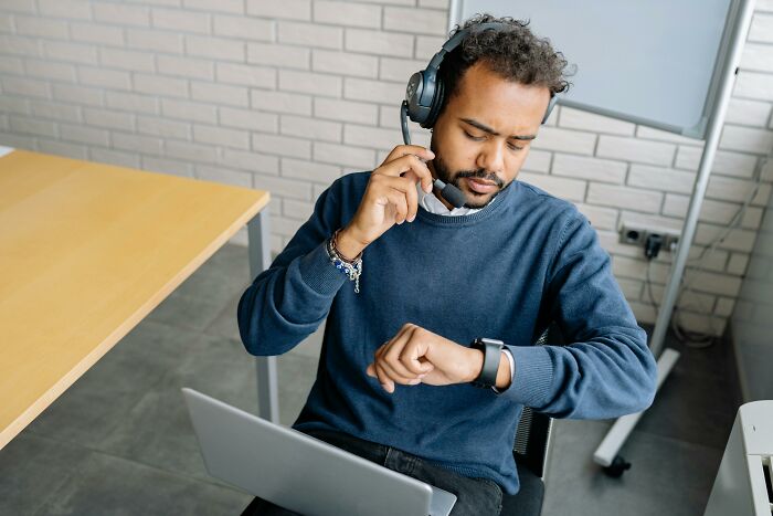 Man wearing a headset checks his watch while seated at a desk with a laptop, illustrating time management habits.