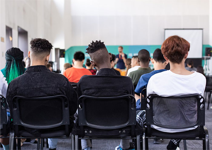 Students seated in a classroom attending a school presentation, viewed from behind in a brightly lit setting.