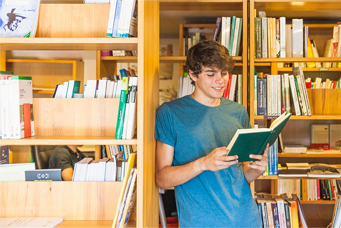 A student in a library, reading a book with shelves full of books in the background, related to school presentations.