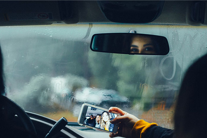 Person taking a selfie in a car on a rainy day, with their reflection visible in the rearview mirror.