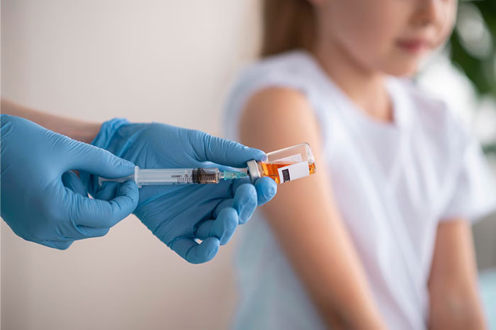 Close-up of gloved hands holding a syringe next to a child in a white shirt, illustrating a school vaccination presentation.