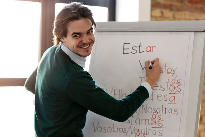 Man smiling during a school presentation, writing on a whiteboard with Spanish verbs.