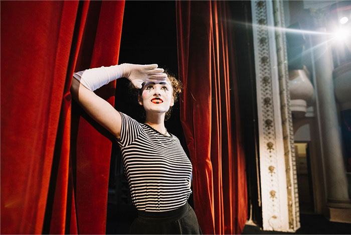 Mime artist on stage in a striped shirt, performing in front of a red curtain during a school presentation.