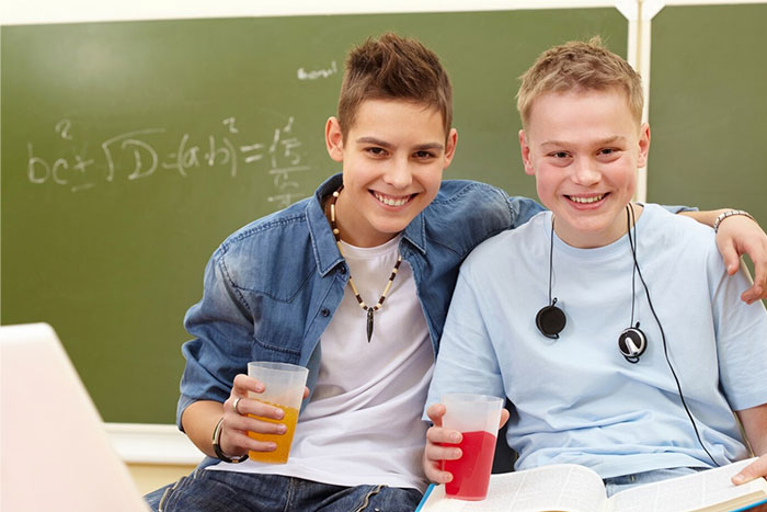 Two students smiling in front of a chalkboard, one holding a drink, representing a school presentation setting.