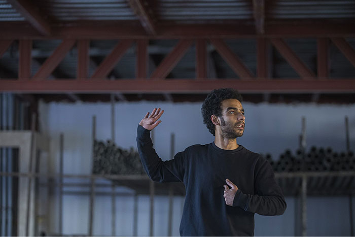 Person gesturing during a school presentation in a large room, wearing a dark sweater.