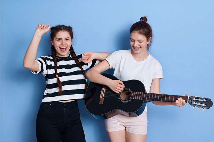 Two girls in a school presentation; one holds a guitar, both smiling against a blue background.