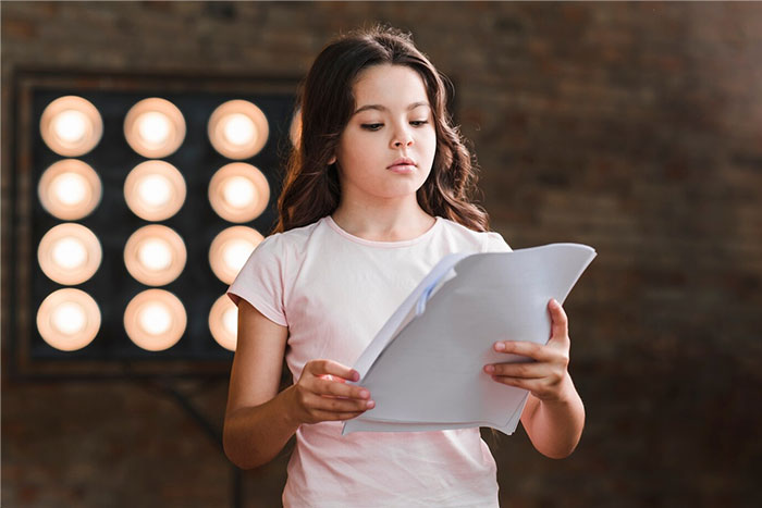 Young girl holding papers, preparing for a school presentation, standing in front of bright lights.