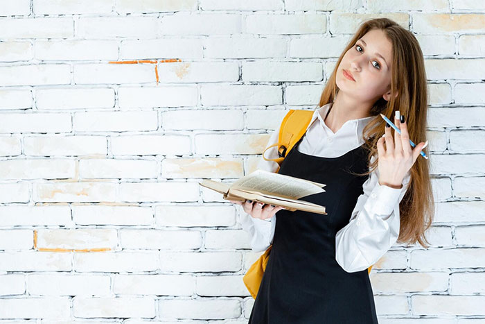 Student with book and backpack stands against brick wall, illustrating school presentations.