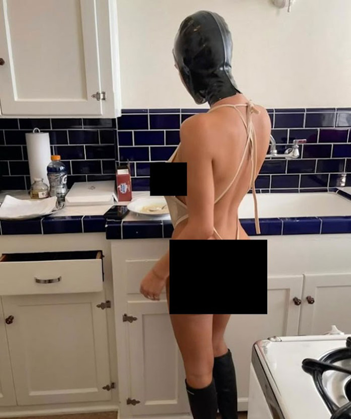 Bianca Censori in a kitchen wearing minimal clothing and a black head covering, standing by a counter.