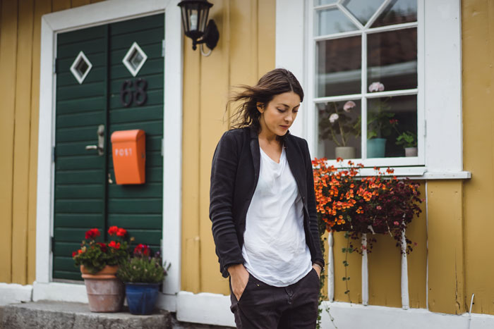 Woman pondering outside a house with hands in pockets, questioning boyfriend's refusal to invite her over.