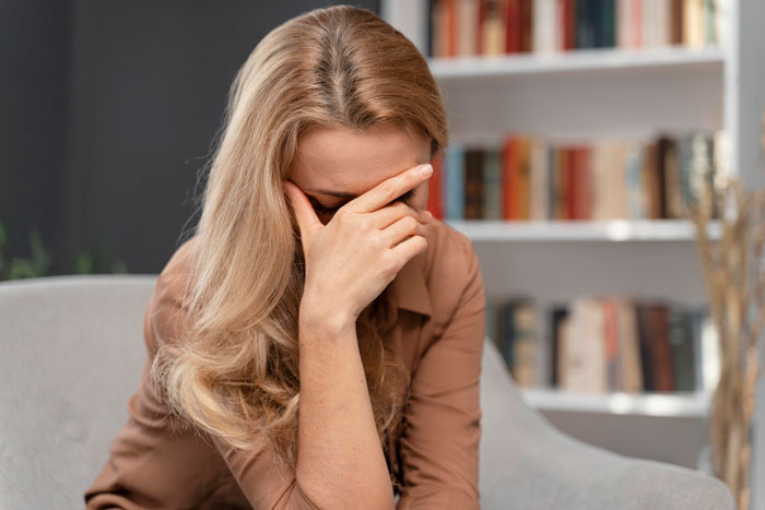 Woman looking puzzled, sitting in a cozy room with bookshelves, wondering about her boyfriend's refusal to let her visit his home.