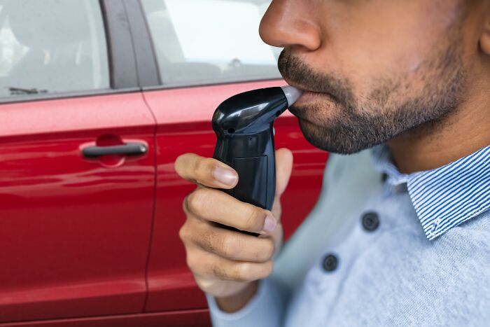 Traveler using breathalyzer next to a red car, capturing a cringeworthy travel moment.