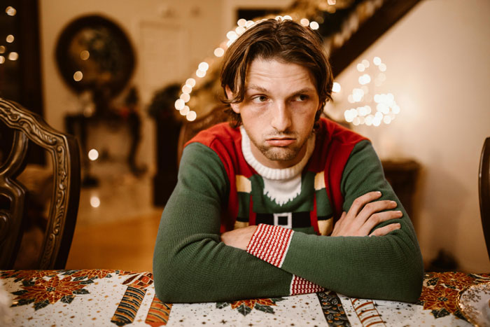 Man in a festive sweater looking pensive at a Christmas dinner table with holiday decorations in the background.