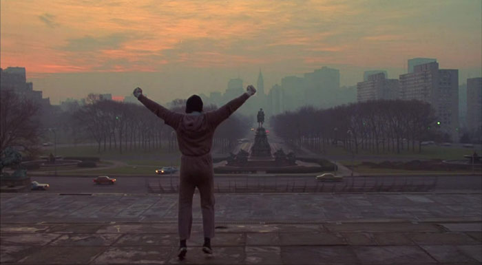 Person posing on steps in a triumphant stance at sunrise, reminiscent of famous Rocky scenes from movies.