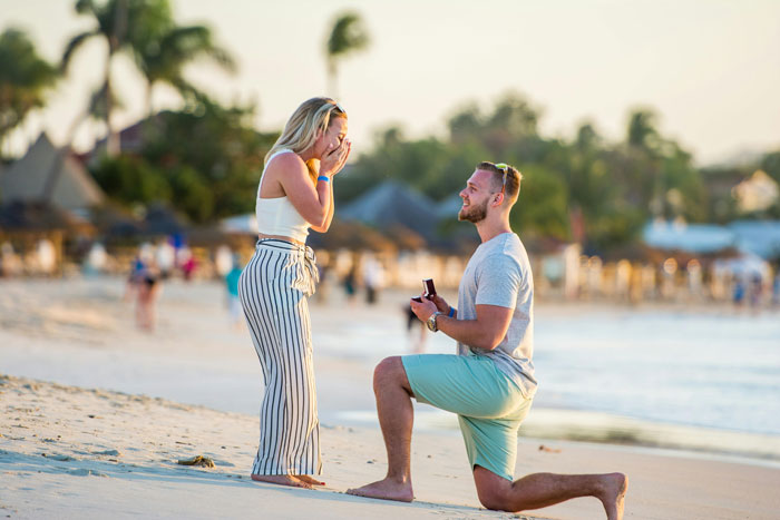 Man proposing on beach during brother's wedding engagement, holding ring box as woman reacts.