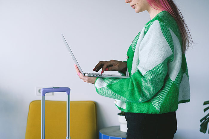 Woman in green sweater using a laptop, standing next to a purple suitcase, addressing wedding chores.