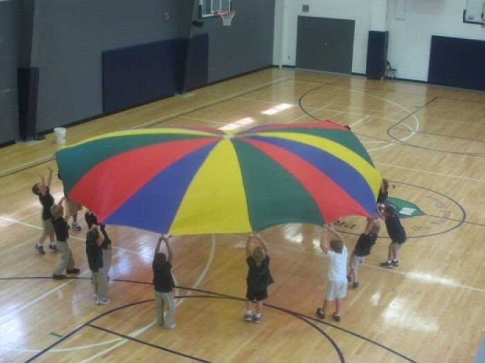 Children playing with a colorful parachute during an 80s gym fitness class in a school gymnasium.