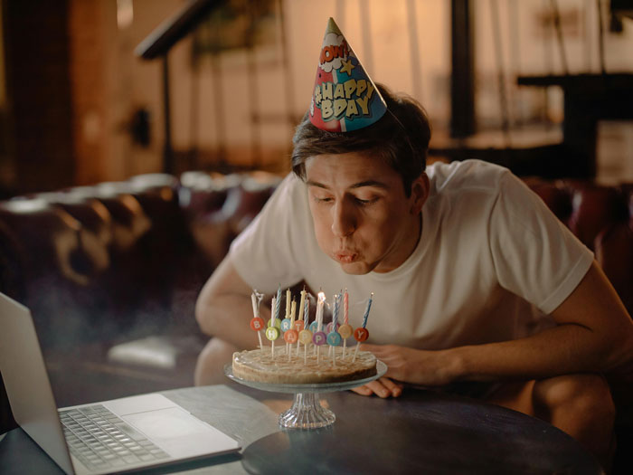 Young man at a birthday party blowing out candles on a cake, wearing a party hat.