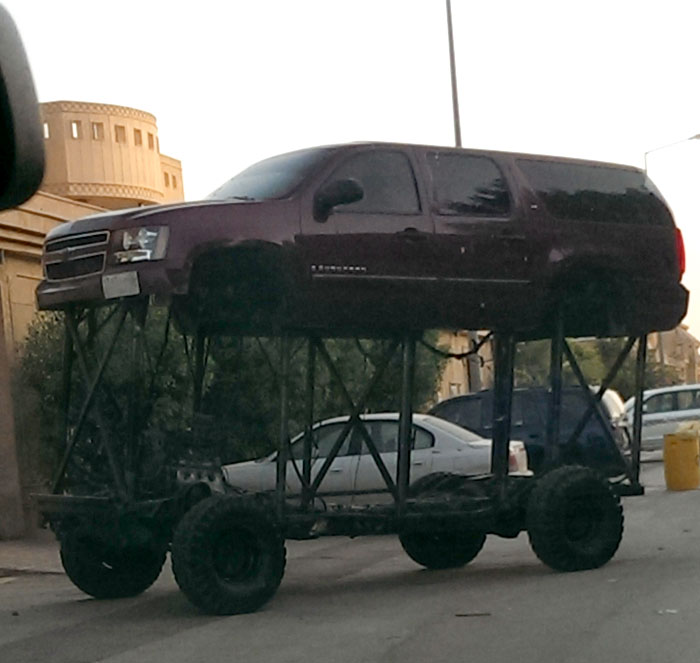 Lifted SUV on jacked-up platform, illustrating a car-fails scenario on a street with buildings in the background.