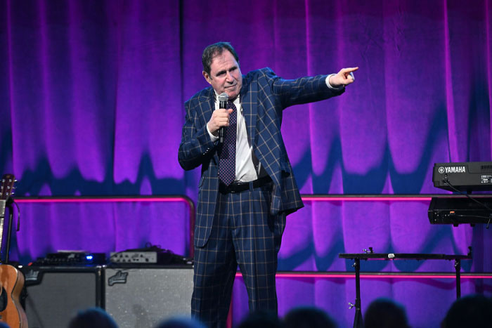 A-List star from Only Murders In The Building Season 4, in a plaid suit, performing on stage with a microphone.