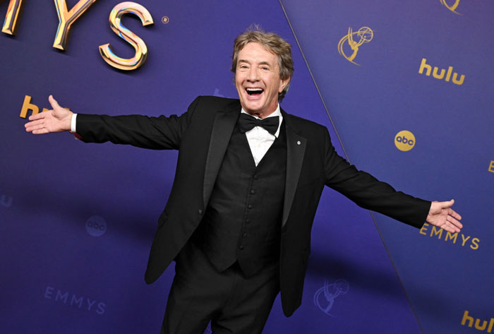 A cast member of "Only Murders In The Building" in a black tuxedo, posing at an awards event with arms outstretched.