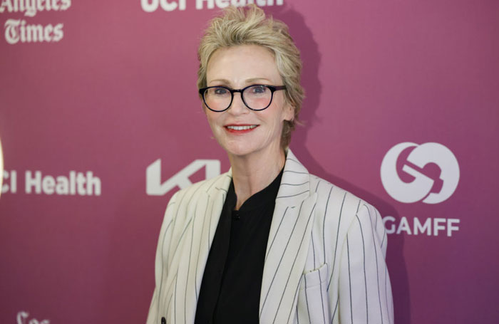 A cast member at a red carpet event, wearing glasses and a striped blazer, smiling against a purple backdrop.