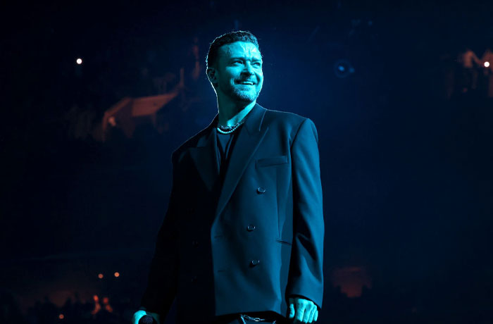 Musician in a dark jacket performing on stage with dramatic blue lighting, part of Trolls 3 musical cast.