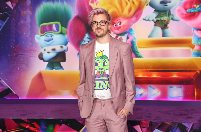 A person in a pink suit poses at the Trolls 3 premiere, with colorful characters in the background.