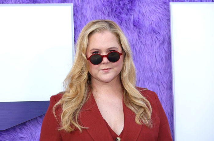 Person in a red outfit and sunglasses at Trolls 3 musical event against purple backdrop.