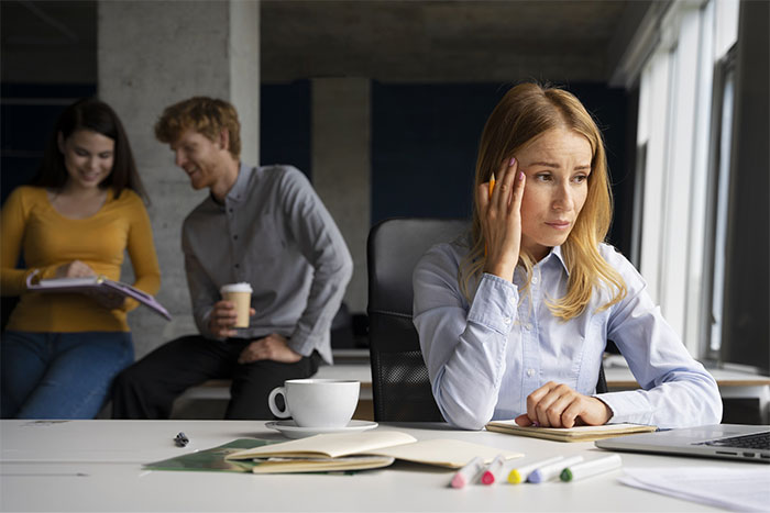 A worried woman at a desk, with a notebook and coffee, while two colleagues chat in the background.