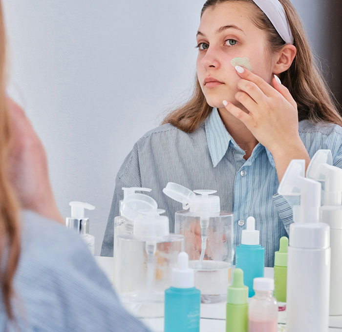 Teen using skincare products in front of a mirror, surrounded by various bottles.