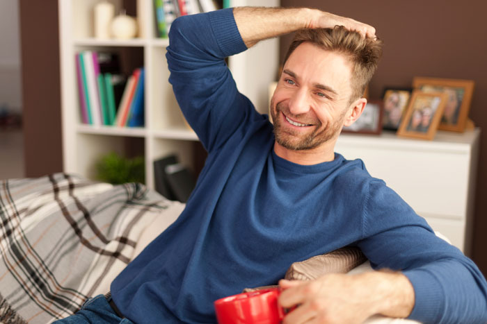 Man in blue sweater, smiling while holding a red mug, sitting on a couch. Shelves with books and photos in the background.