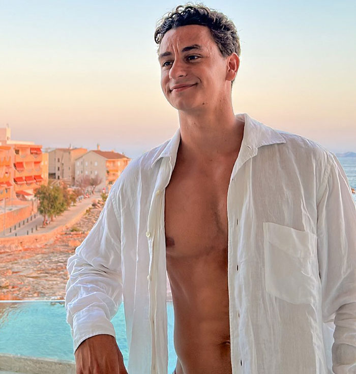 Young man in an open white shirt smiling on a balcony, with a sunset view over buildings and sea.