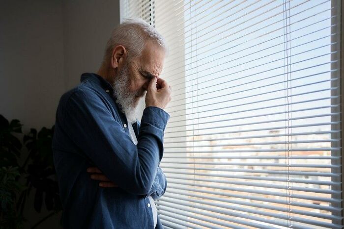 Elderly man in a blue shirt, looking stressed by a window, reflecting on challenges of aging.