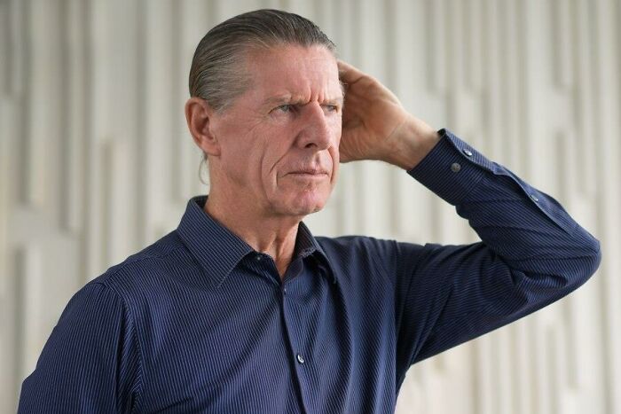 Man with hand on head, wearing a blue shirt, reflecting on unexpected challenges of aging.