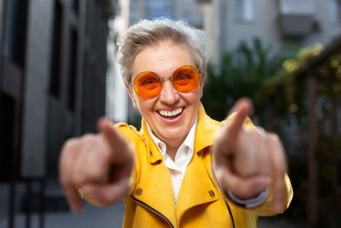 Smiling person in yellow jacket and orange sunglasses pointing, symbolizing unexpected aspects of aging.