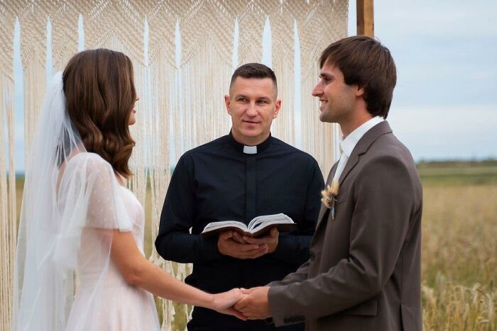 Bride and groom holding hands at an outdoor wedding ceremony, with officiant reading from a book.