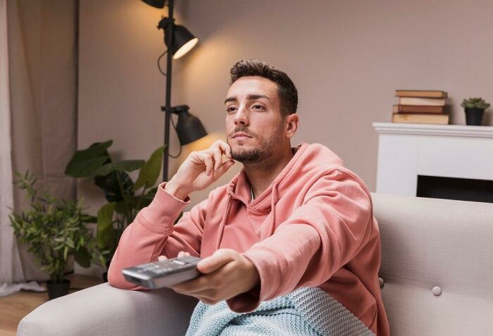 Man in a pink hoodie, sitting on a couch, holding a remote and contemplating, reflecting the theme of thoughts on his mind.