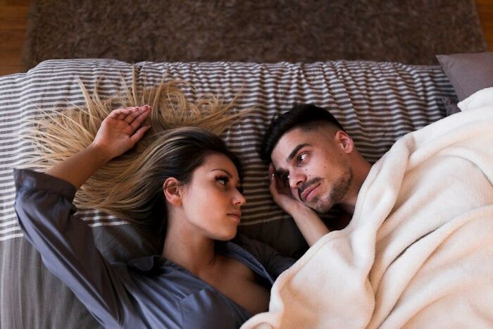 Couple lying in bed, woman looking curiously at man, pondering thoughts.