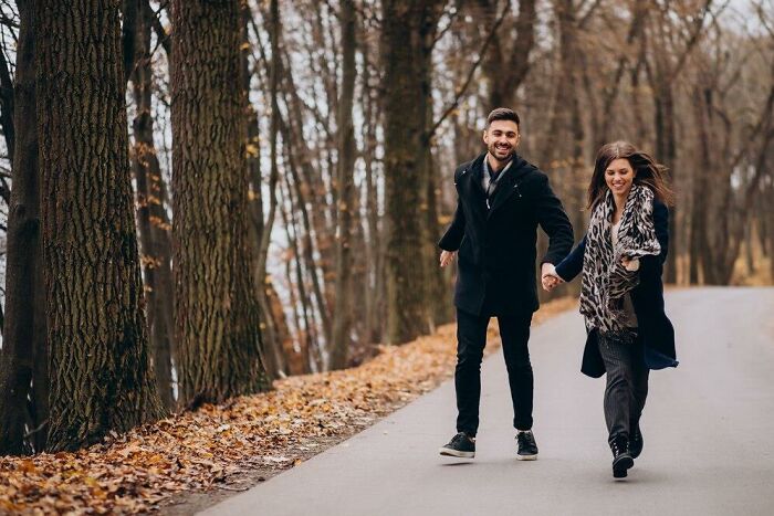 A couple happily walking through a scenic autumn path, surrounded by tall trees and fallen leaves.