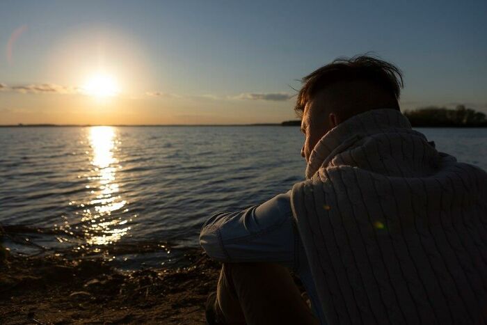 Man thinking while watching sunset over a calm lake, wrapped in a blanket.