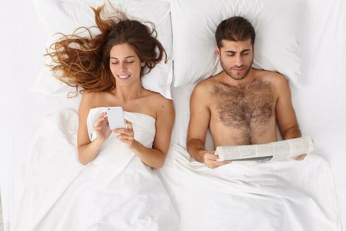 Man reading a newspaper and woman using a phone in bed, illustrating curiosity about men's thoughts.