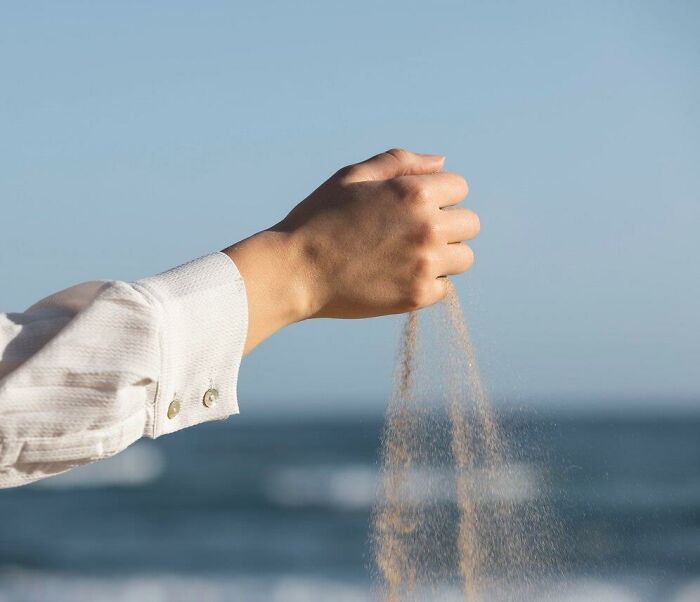 Hand in white sleeve holding sand, symbolizing thoughts drifting away, against a blurred ocean background.